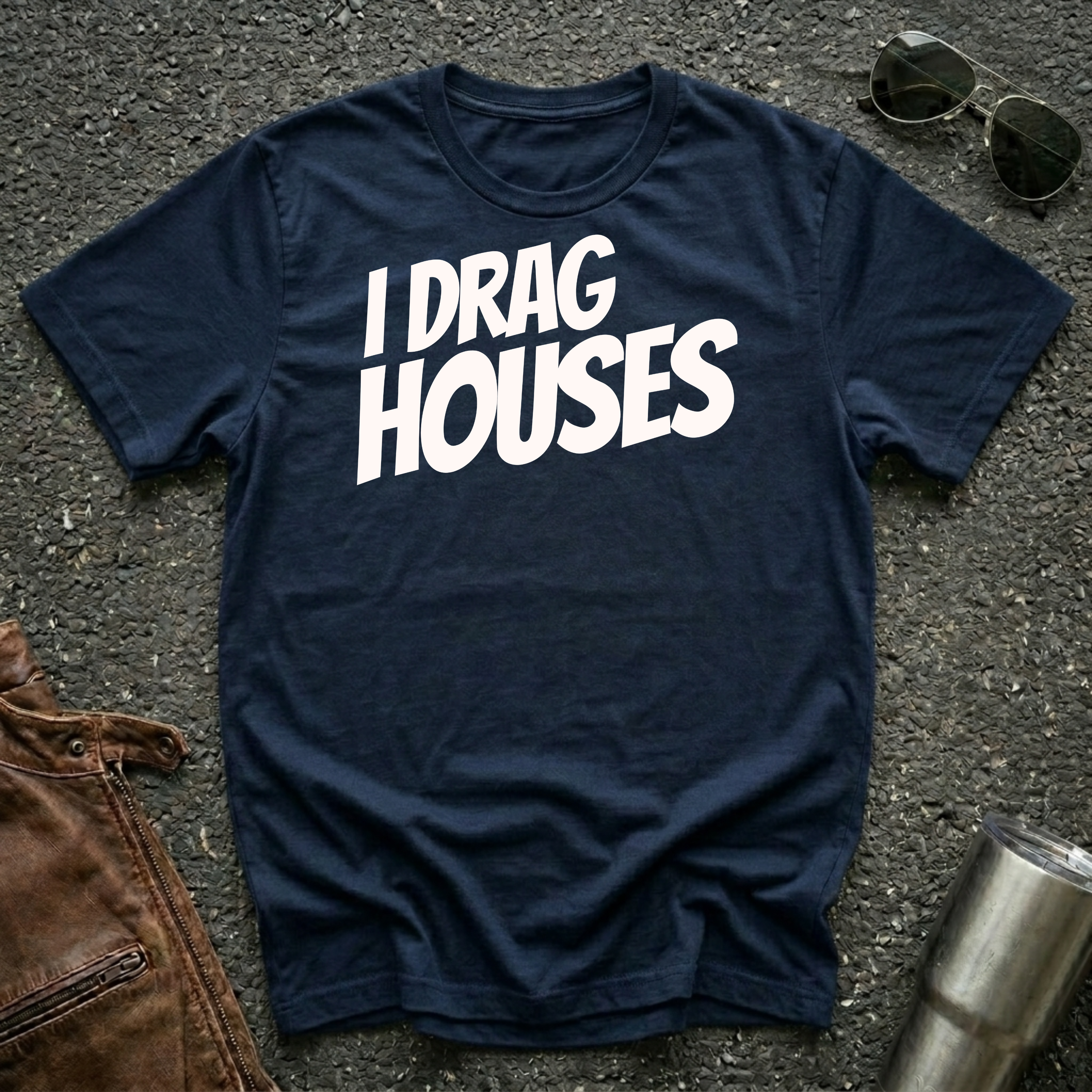 A navy short-sleeve t-shirt with the phrase 'I DRAG HOUSES' printed in bold  letters across the chest, laid flat on a dark asphalt-like surface with sunglasses, brown leather jacket and a metal tumbler nearby.