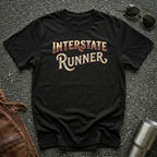 A black short-sleeve graphic T-shirt laid flat on a dark textured surface, with the words "INTERSTATE RUNNER" printed across the chest in a distressed cream and maroon font. Sunglasses, part of a brown leather jacket, and a metal tumbler are placed around it as styling props.