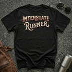 A black short-sleeve graphic T-shirt laid flat on a dark textured surface, with the words "INTERSTATE RUNNER" printed across the chest in a distressed cream and maroon font. Sunglasses, part of a brown leather jacket, and a metal tumbler are placed around it as styling props.