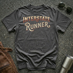 A dark heather gray graphic t-shirt with 'INTERSTATE RUNNER' printed in a distressed maroon and cream retro font, laid flat on a textured gray asphalt background with sunglasses, brown leather jacket  and a metal tumbler as props.