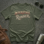 A military green short-sleeve graphic T-shirt laid flat on a dark textured surface, with the words "INTERSTATE RUNNER" printed across the chest in a distressed cream and maroon font. Sunglasses, part of a brown leather jacket, and a metal tumbler are placed around it as styling props.