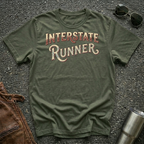 An olive green crewneck t-shirt with 'INTERSTATE RUNNER' printed in a retro cream and maroon font, laid flat on a dark asphalt surface with sunglasses, a brown leather jacket, and a metal tumbler as styling props.