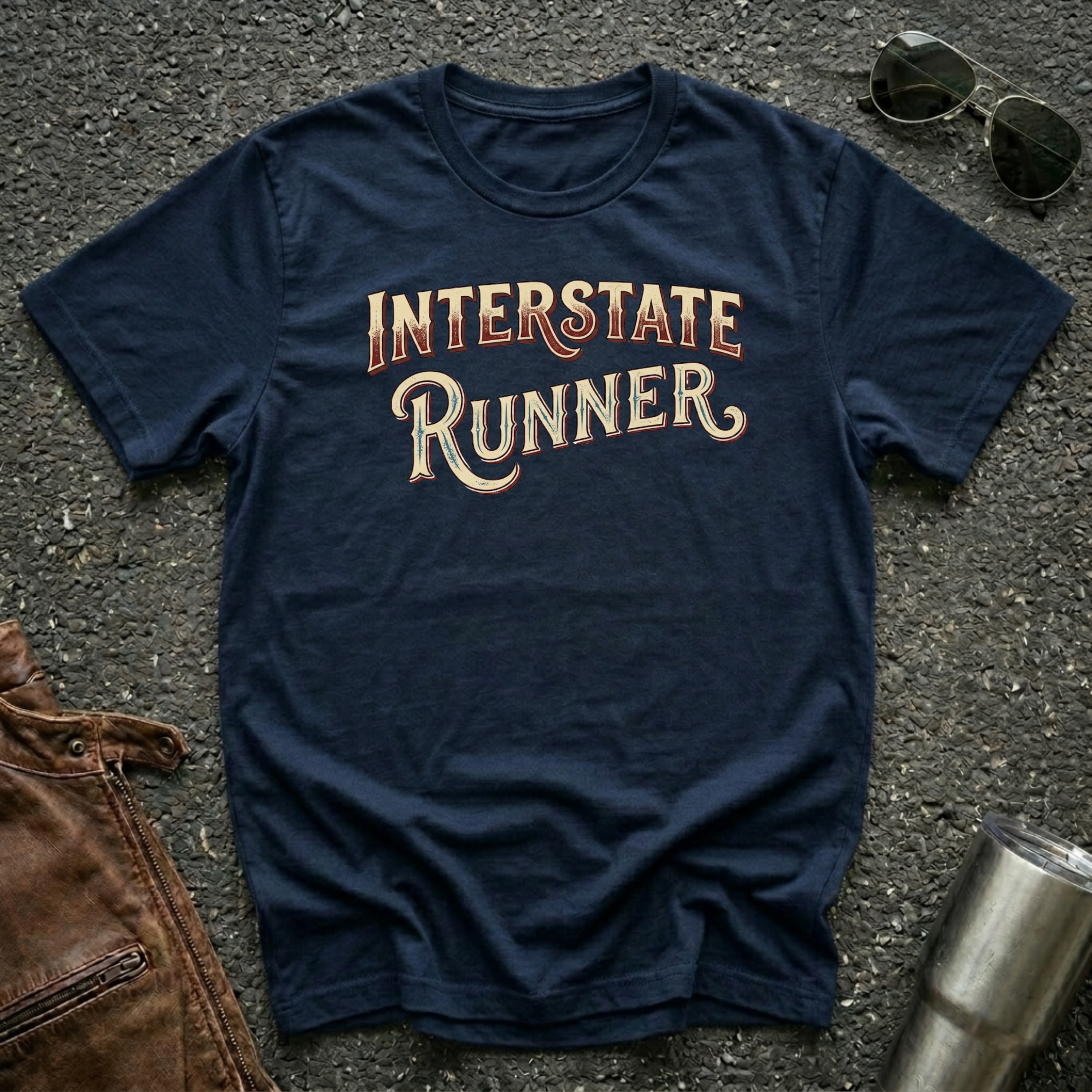 A navy short-sleeve graphic T-shirt laid flat on a dark textured surface, with the words "INTERSTATE RUNNER" printed across the chest in a distressed cream and maroon font. Sunglasses, part of a brown leather jacket, and a metal tumbler are placed around it as styling props.