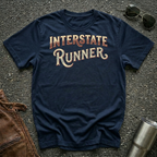 A navy blue graphic t-shirt with 'INTERSTATE RUNNER' printed in a distressed cream and maroon font, styled in a flat lay on asphalt with sunglasses, a brown leather jacket, and a metal tumbler.