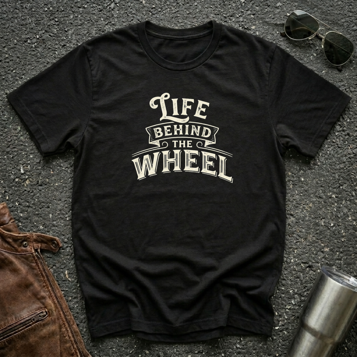 A black short-sleeve t-shirt with a distressed cream graphic reading 'LIFE BEHIND THE WHEEL' laid flat on a dark asphalt-like surface. Sunglasses, brown leather jacket, and a metal tumbler are partially visible around the shirt as styling props.