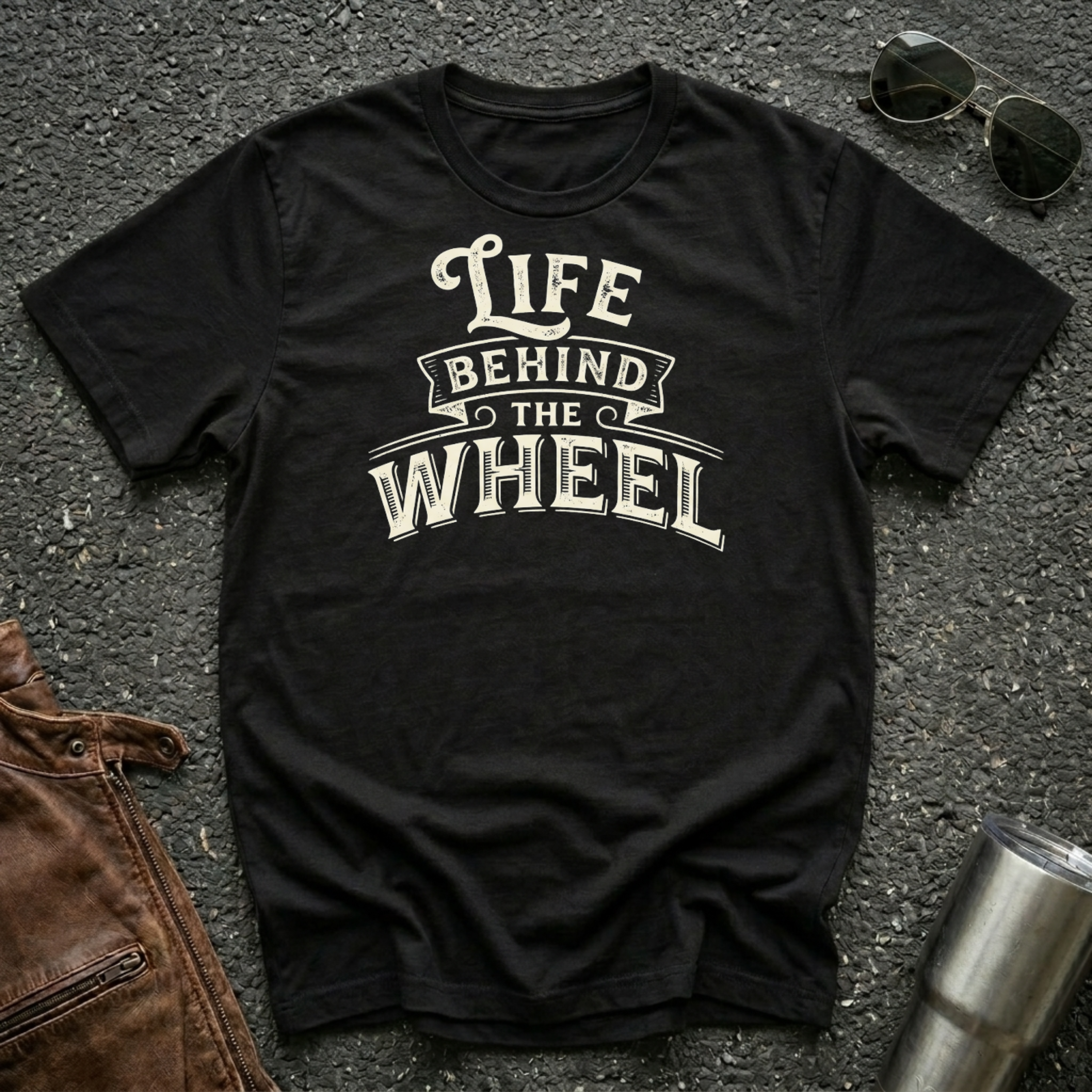 A black short-sleeve t-shirt with a distressed cream graphic reading 'LIFE BEHIND THE WHEEL' laid flat on a dark asphalt-like surface. Sunglasses, brown leather jacket, and a metal tumbler are partially visible around the shirt as styling props.