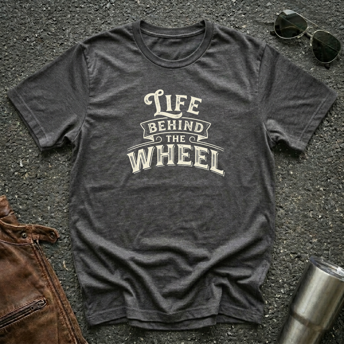 A dark heather short-sleeve t-shirt with a distressed cream graphic reading 'LIFE BEHIND THE WHEEL' laid flat on a dark asphalt-like surface. Sunglasses, brown leather jacket, and a metal tumbler are partially visible around the shirt as styling props.
