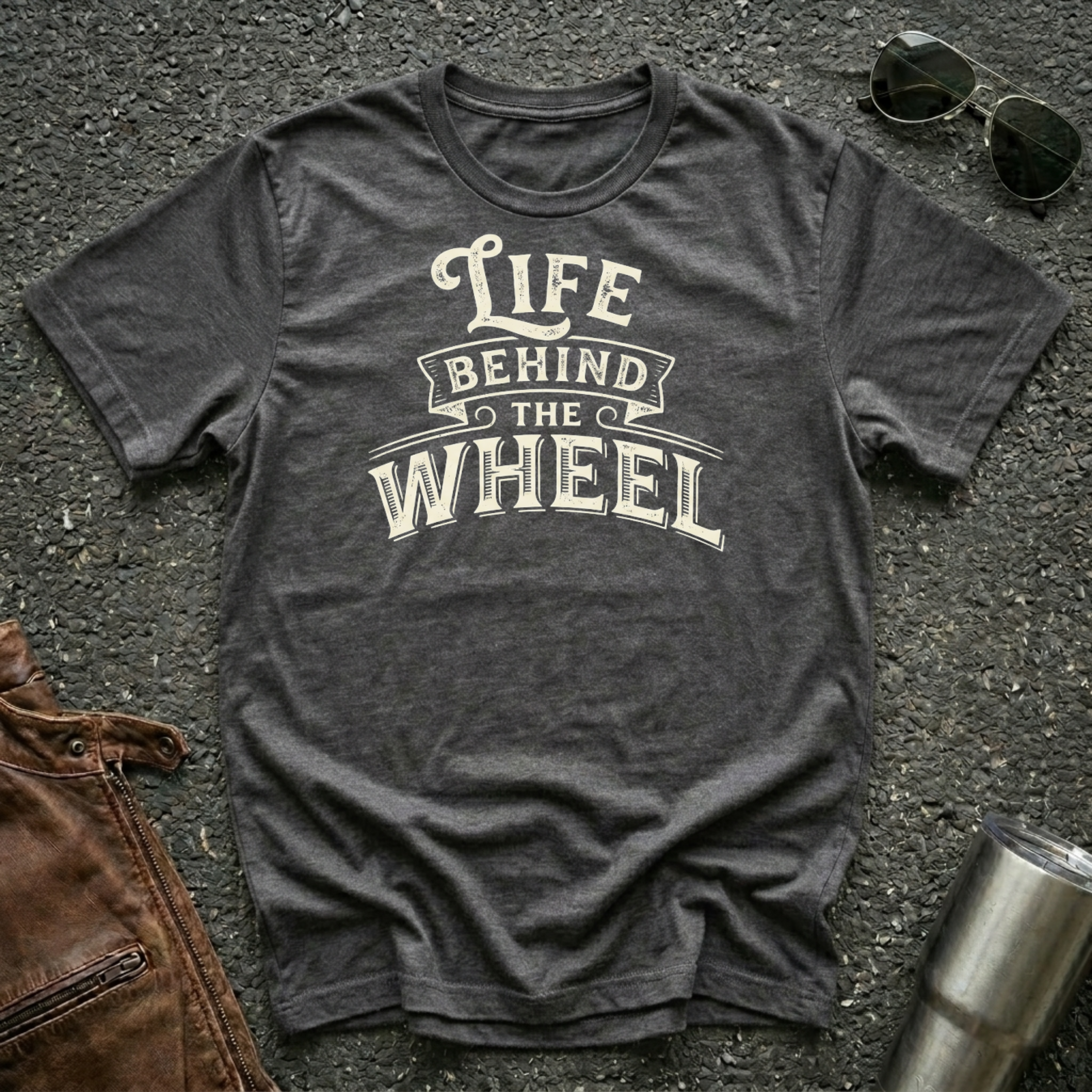 A dark heather short-sleeve t-shirt with a distressed cream graphic reading 'LIFE BEHIND THE WHEEL' laid flat on a dark asphalt-like surface. Sunglasses, brown leather jacket, and a metal tumbler are partially visible around the shirt as styling props.
