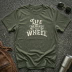 A military green short-sleeve t-shirt with a distressed cream graphic reading 'LIFE BEHIND THE WHEEL' laid flat on a dark asphalt-like surface. Sunglasses, brown leather jacket, and a metal tumbler are partially visible around the shirt as styling props.
