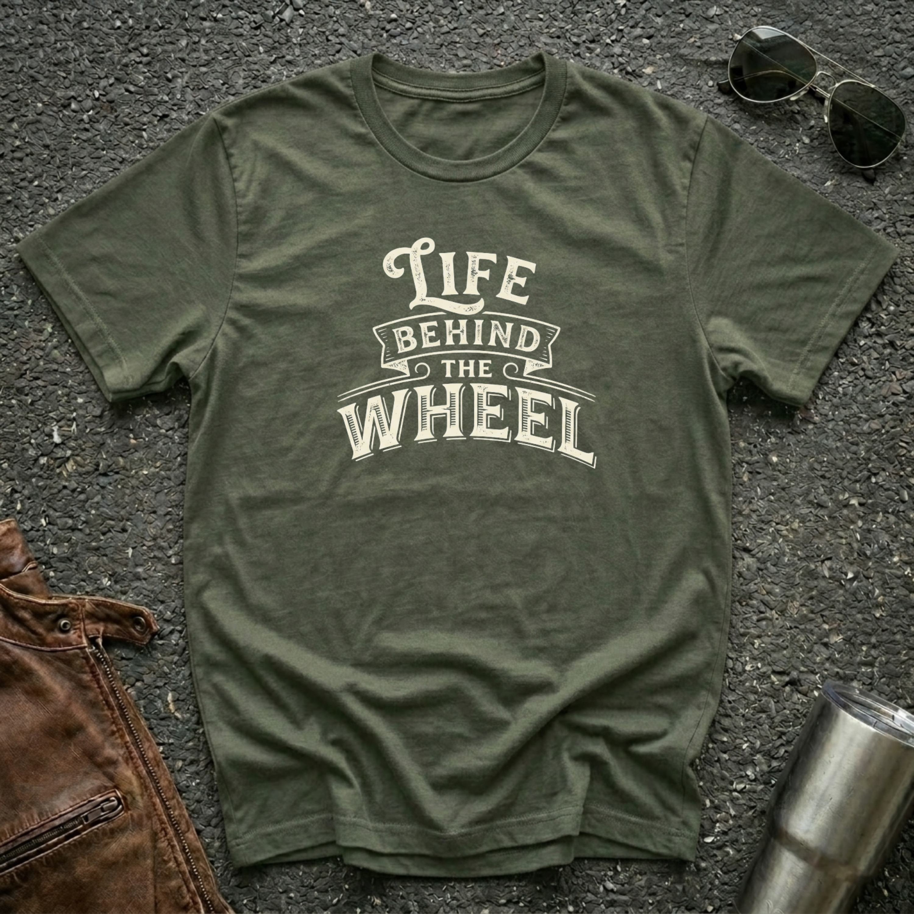 A military green short-sleeve t-shirt with a distressed cream graphic reading 'LIFE BEHIND THE WHEEL' laid flat on a dark asphalt-like surface. Sunglasses, brown leather jacket, and a metal tumbler are partially visible around the shirt as styling props.