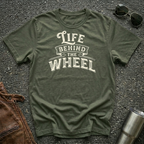 A military green short-sleeve t-shirt with a distressed cream graphic reading 'LIFE BEHIND THE WHEEL' laid flat on a dark asphalt-like surface. Sunglasses, brown leather jacket, and a metal tumbler are partially visible around the shirt as styling props.