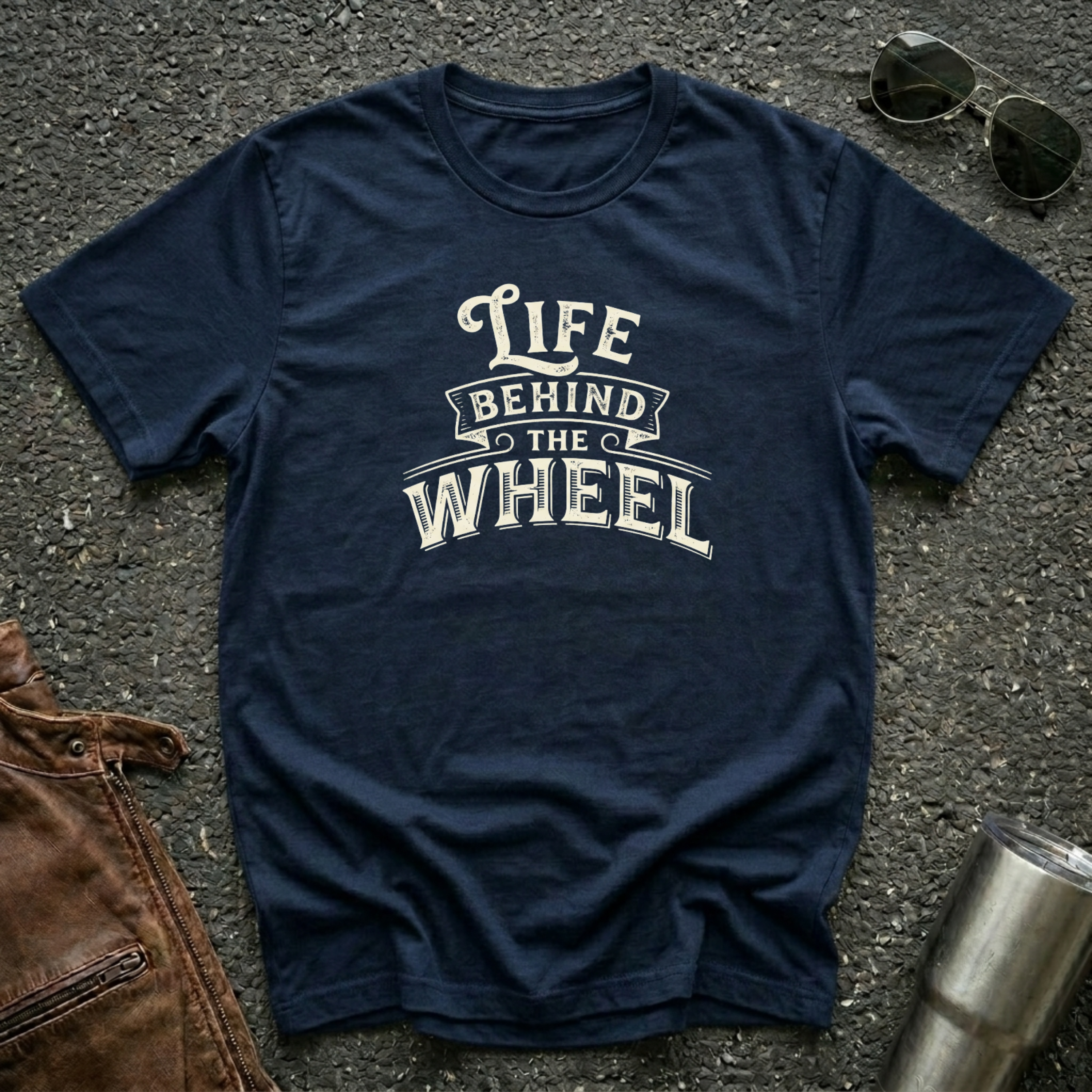 A navy blue short-sleeve t-shirt with a distressed cream graphic reading 'LIFE BEHIND THE WHEEL' laid flat on a dark asphalt-like surface. Sunglasses, brown leather jacket, and a metal tumbler are partially visible around the shirt as styling props.