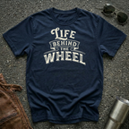 A navy blue short-sleeve t-shirt with a distressed cream graphic reading 'LIFE BEHIND THE WHEEL' laid flat on a dark asphalt-like surface. Sunglasses, brown leather jacket, and a metal tumbler are partially visible around the shirt as styling props.