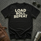 A black short-sleeve t-shirt with the words 'LOAD ROLL REPEAT' printed in distressed cream lettering across the chest, laid flat on a dark asphalt-like surface with sunglasses, brown leather jacket, and a metal tumbler nearby.