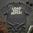 A dark heather gray t-shirt with the words 'LOAD ROLL REPEAT' printed in distressed cream lettering, laid flat on a dark asphalt surface with sunglasses, a brown leather jacket, and a metal tumbler nearby.