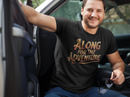 A man sitting in the driver's seat of a truck wearing a black t-shirt with a gold graphic that reads 'ALONG FOR THE ADVENTURE'.