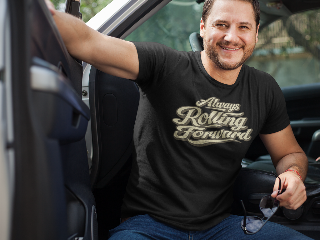 A smiling man sitting in the driver's seat of a truck wearing a black t-shirt with a metallic gold script graphic that reads 'Always Rolling Forward'.