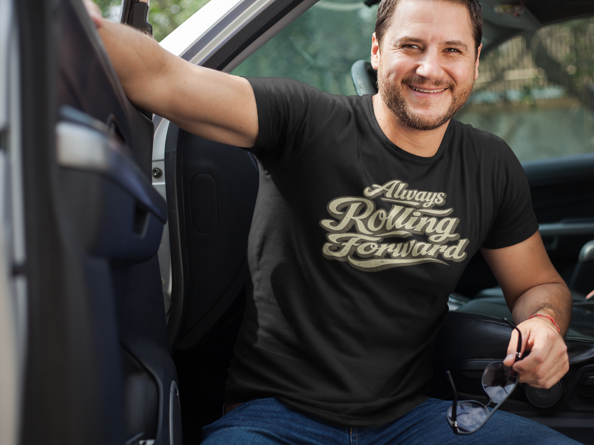 A smiling man sitting in the driver's seat of a truck wearing a black t-shirt with a metallic gold script graphic that reads 'Always Rolling Forward'.