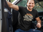 A smiling man sitting in the driver's seat of a truck wearing a black graphic t-shirt with a retro-style design that reads 'ARE WE THERE YET? ASKING FOR THE 47TH TIME'.