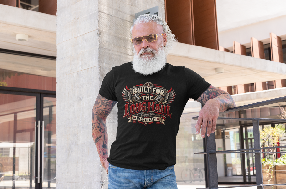 A bearded man with tattoos and a white wig leans against a concrete pillar in an outdoor urban setting, wearing a black graphic t-shirt with a red and gold 'Built for the Long Haul Made to Last' design.