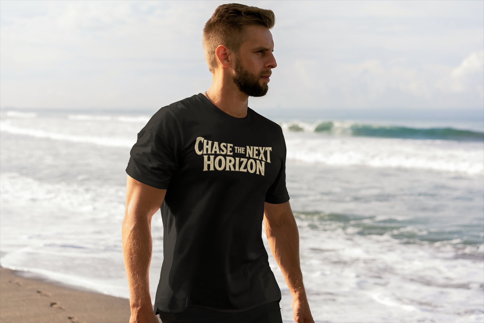 A man standing on a beach wearing a black t-shirt with the text 'Chase the Next Horizon' printed in a gold serif font on the chest.