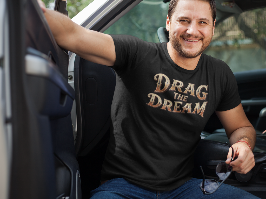 A smiling man sitting in a truck wearing a black t-shirt with the text 'DRAG THE DREAM' in a metallic gold and bronze gradient font. He is wearing blue jeans and holding sunglasses.