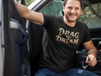 A smiling man sitting in a truck wearing a black t-shirt with the text 'DRAG THE DREAM' in a metallic gold and bronze gradient font. He is wearing blue jeans and holding sunglasses.