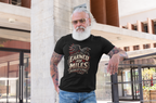 A man with a white beard, sunglasses, and tattoos leans against a modern concrete pillar, wearing a black graphic t-shirt that reads 'Earned My Miles Nothing Left To Prove' with a red raven design.
