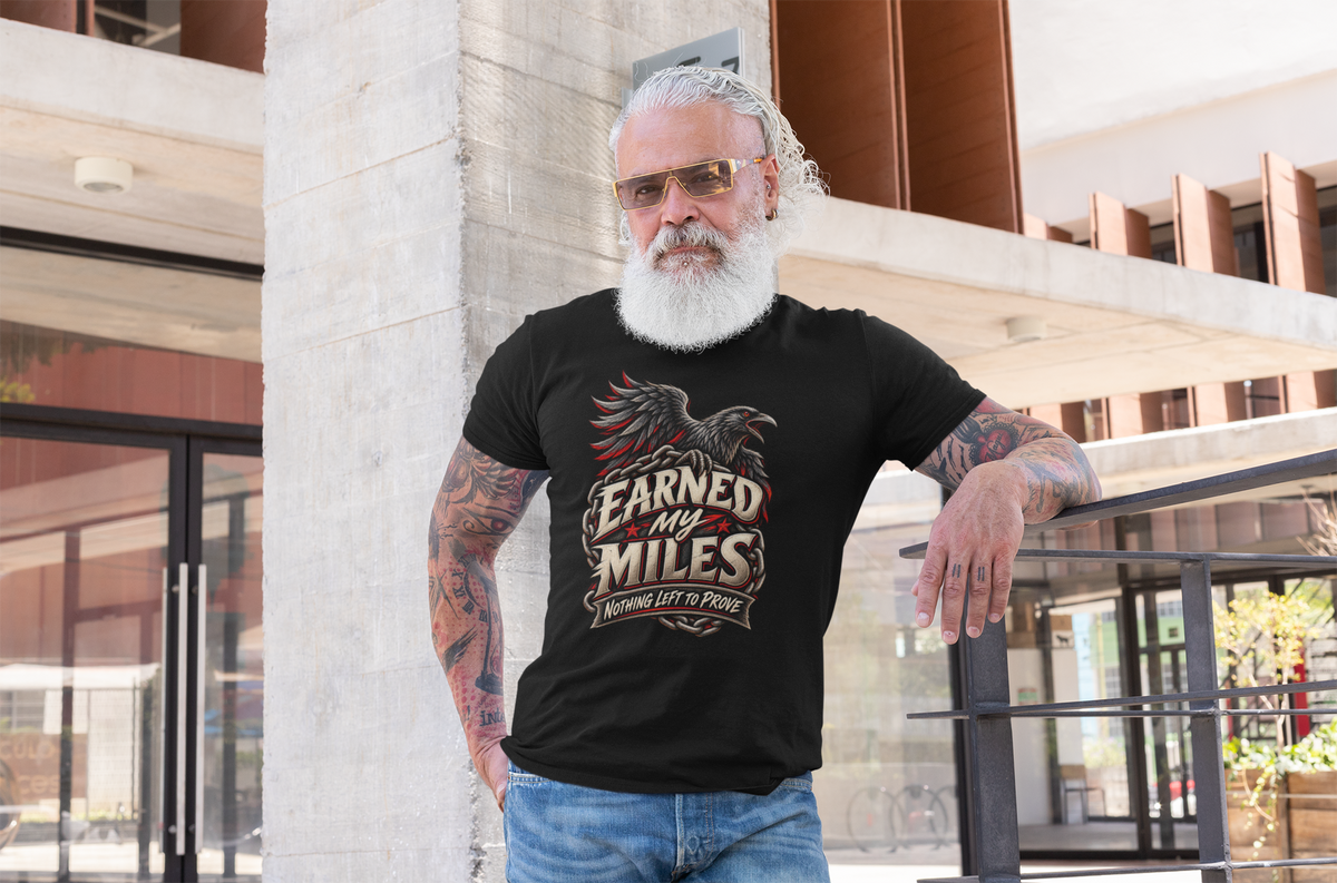 A man with a white beard, sunglasses, and tattoos leans against a modern concrete pillar, wearing a black graphic t-shirt that reads 'Earned My Miles Nothing Left To Prove' with a red raven design.