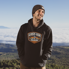 A man wearing a black hoodie with a 'Lost-Ish Again' graphic on the chest and a brown knit beanie, standing outdoors with a mountain and cloud landscape in the background.