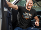 A smiling man sitting in a truck wearing a black graphic t-shirt with the text 'LOST-ISH AGAIN' and a graphic of a man and a dog. He is wearing blue jeans and holding sunglasses.