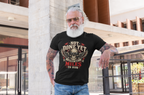 A man with a white beard and sunglasses leans against a concrete pillar in an outdoor urban setting, wearing a black graphic t-shirt with a skull and wings design that reads 'NOT DONE YET STILL GOT MILES TO RUN'.