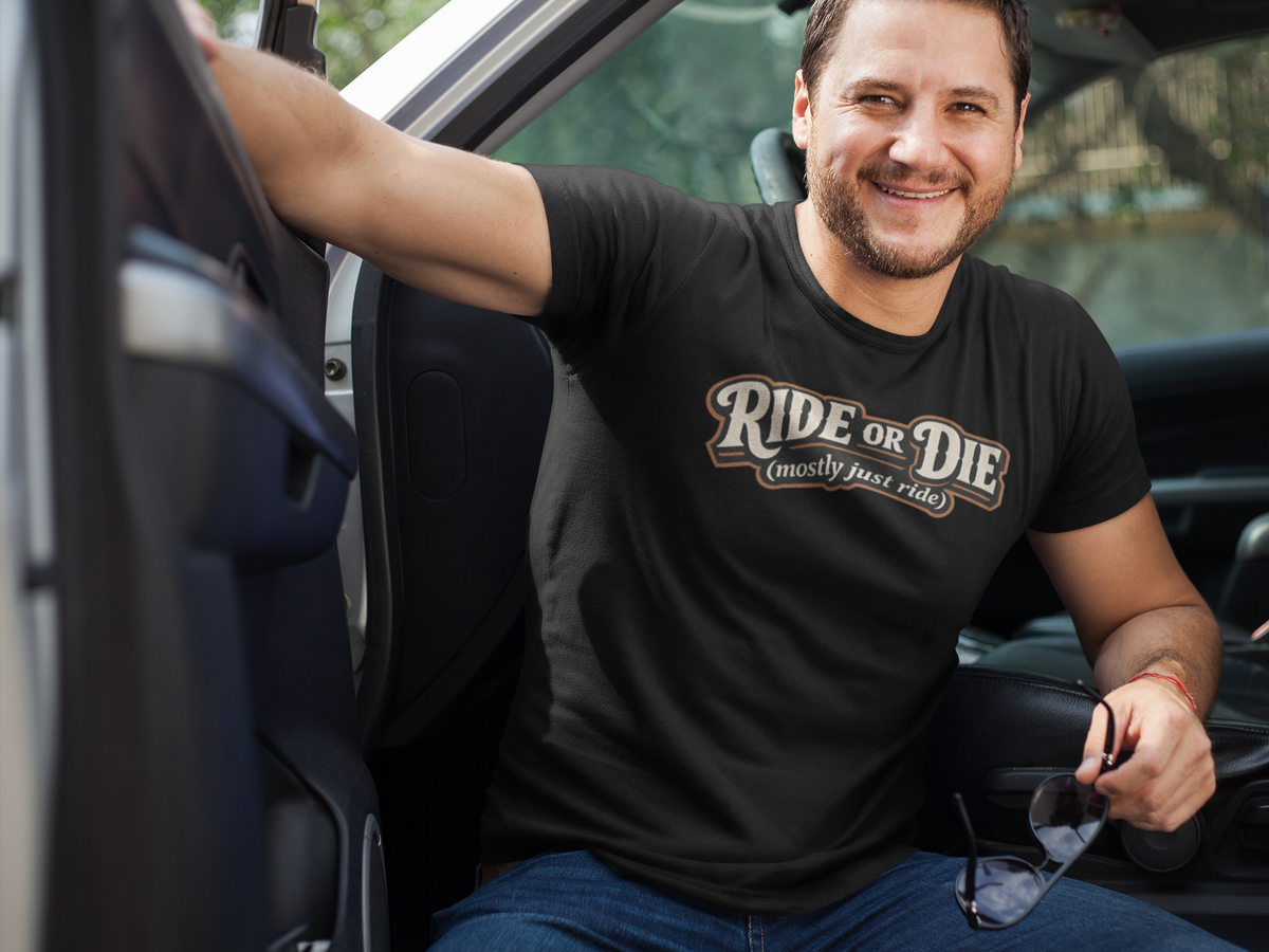 A smiling man sitting in a truck wearing a black graphic t-shirt with the text 'RIDE OR DIE (mostly just ride)' in brown and beige. He is wearing blue jeans and holding sunglasses.