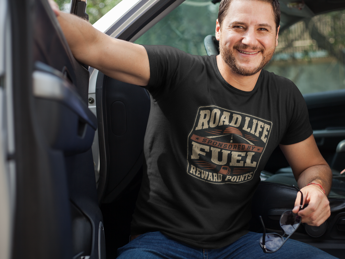A smiling man sitting in a truck wearing a black graphic t-shirt with a distressed design that reads 'ROAD LIFE SPONSORED BY FUEL REWARD POINTS'.