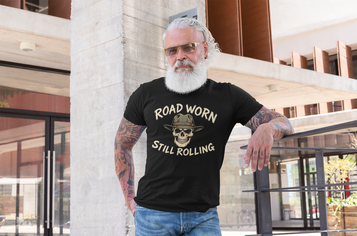 A man with a white beard and sunglasses leans against a concrete pillar in an outdoor urban setting, wearing a black graphic t-shirt with the text 'ROAD WORN STILL ROLLING' and a skull wearing a hat design.