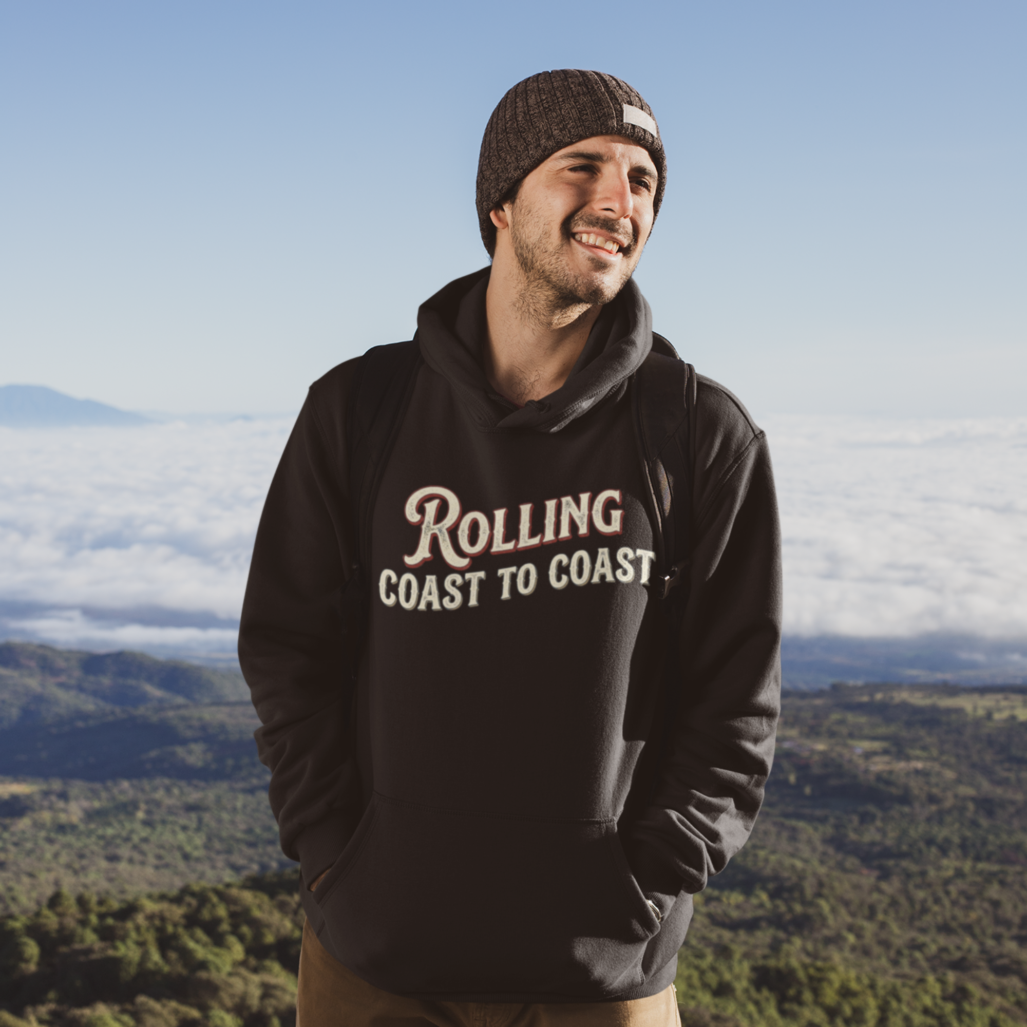 A man wearing a black hoodie with 'Rolling Coast to Coast' printed in beige and white text, standing outdoors against a scenic mountain and cloud background.