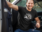 A smiling man sitting in a truckwearing a black crewneck t-shirt with a circular white graphic that reads 'THE RIVERSTONE LIFE Original'.