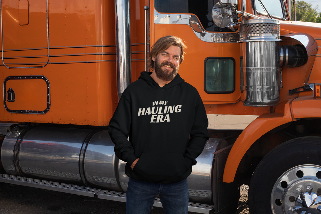 A man with a beard wearing a black hoodie with the text 'IN MY HAULING ERA' in white, standing in front of an orange semi-truck.