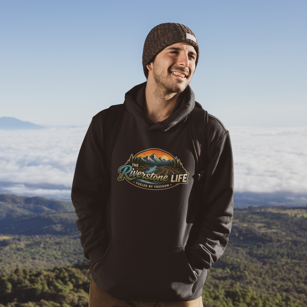 A man wearing a black pullover hoodie with a colorful 'The Riverstone Life- Fueled by Freedom' mountain graphic on the chest, standing outdoors on a mountain peak above a sea of clouds.