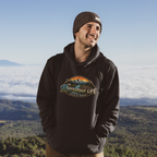 A man wearing a black pullover hoodie with a colorful 'The Riverstone Life- Fueled by Freedom' mountain graphic on the chest, standing outdoors on a mountain peak above a sea of clouds.