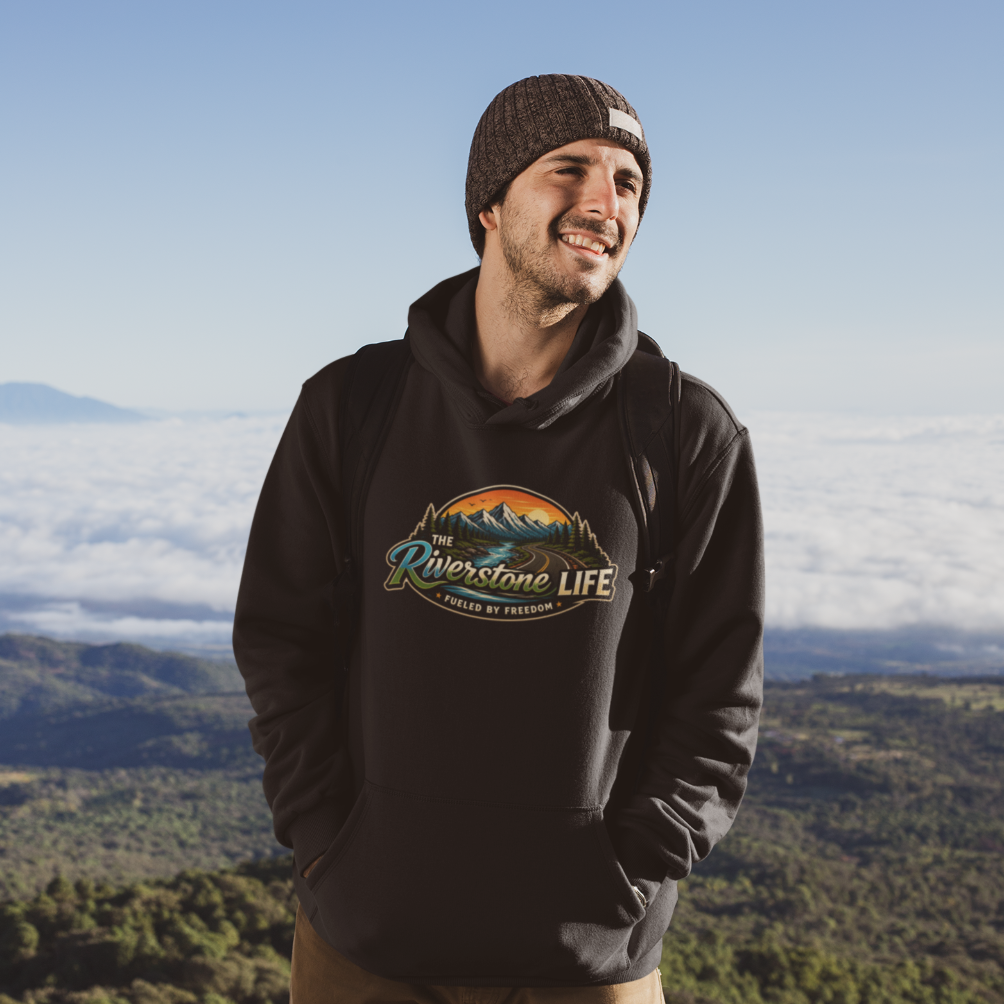 A man wearing a black pullover hoodie with a colorful 'The Riverstone Life- Fueled by Freedom' mountain graphic on the chest, standing outdoors on a mountain peak above a sea of clouds.