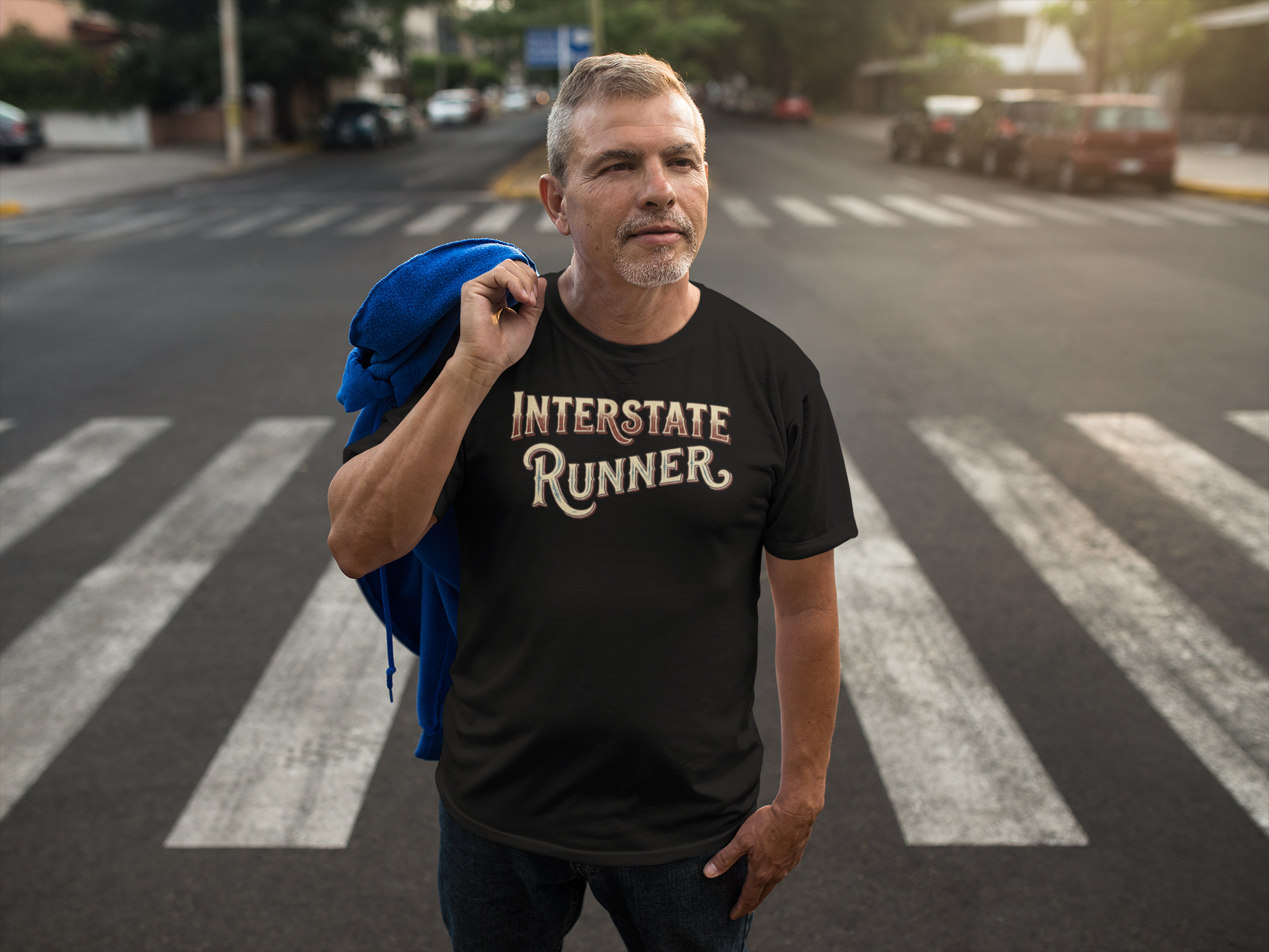 A man standing on a city crosswalk wearing a black graphic t-shirt with 'INTERSTATE RUNNER' printed in a stylized beige and gold font. He has a blue jacket draped over his shoulder, with a blurred street background.