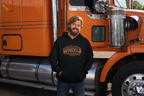 A man with a beard wearing a black hoodie with the text 'KEEP THEY WHEELS WHEELING' stands in front of an orange semi-truck.