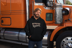 A man with a beard wearing a black hoodie with 'Interstate Runner' printed on the chest, standing in front of an orange semi-truck.
