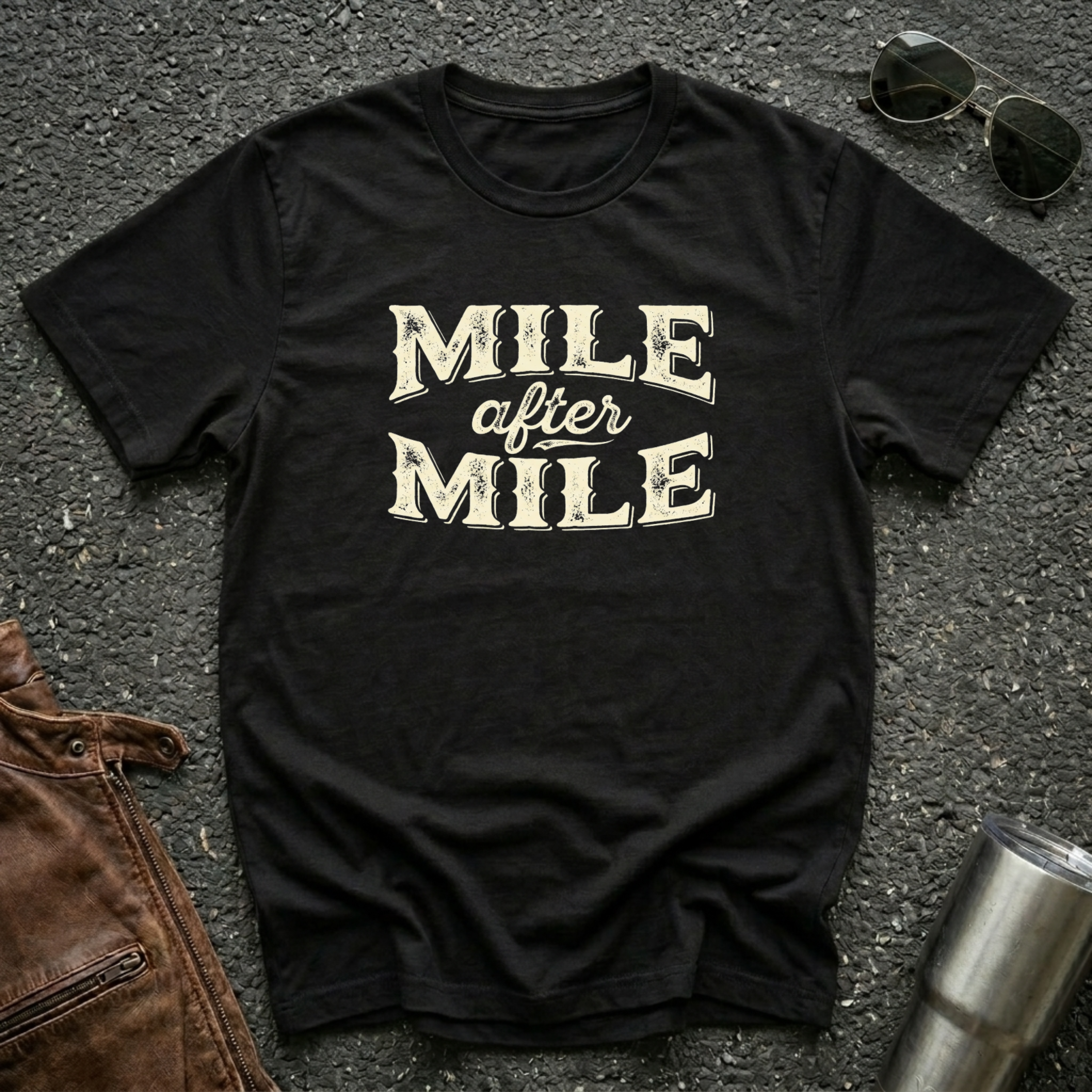 A black graphic t-shirt laid flat on asphalt with sunglasses, a brown leather jacket, and a metal tumbler nearby. The shirt features the text 'MILE after MILE' in distressed cream lettering.