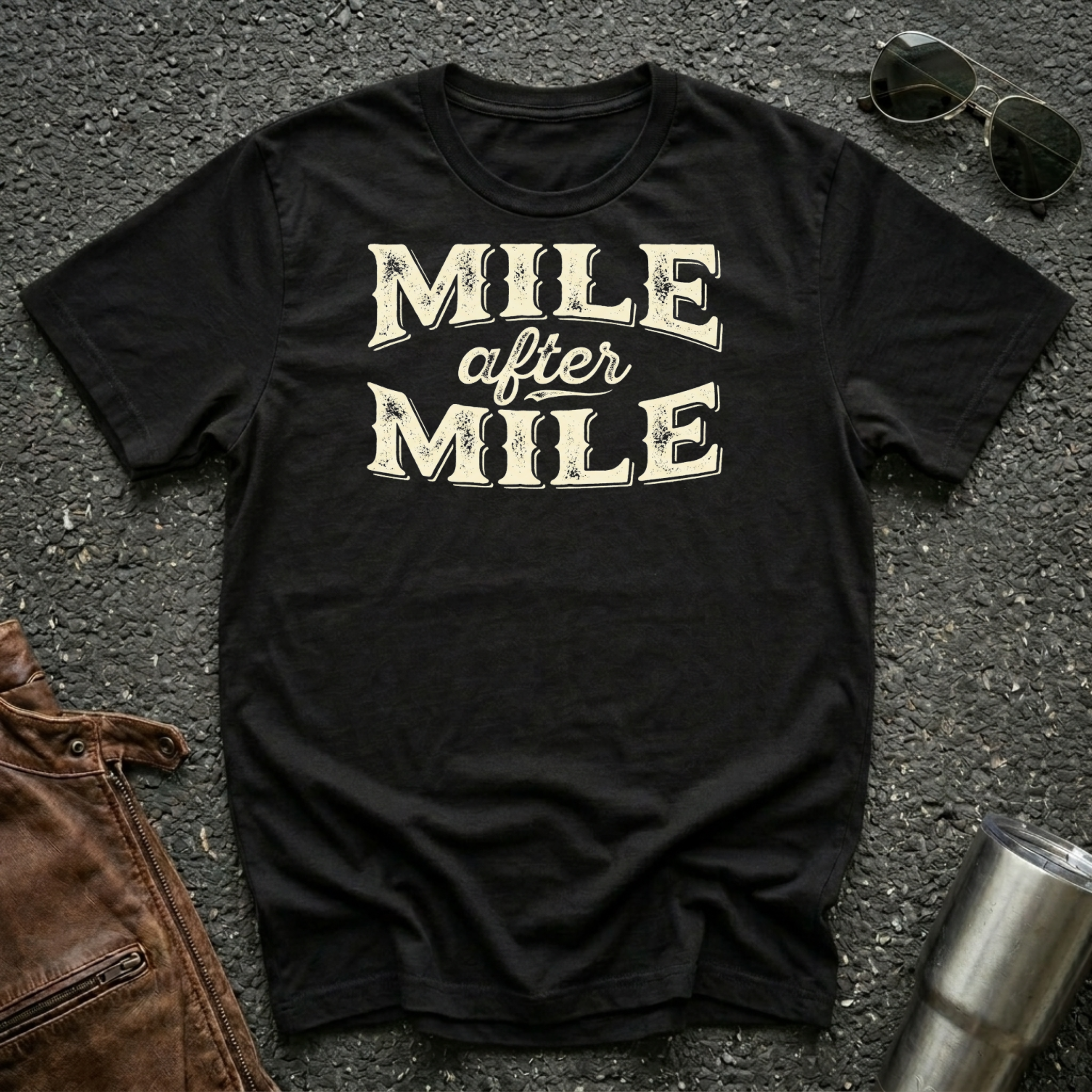 A black graphic t-shirt laid flat on asphalt with sunglasses, a brown leather jacket, and a metal tumbler nearby. The shirt features the text 'MILE after MILE' in distressed cream lettering.