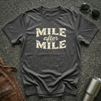 A dark heather graphic t-shirt laid flat on asphalt with sunglasses, a brown leather jacket, and a metal tumbler nearby. The shirt features the text 'MILE after MILE' in distressed cream lettering.