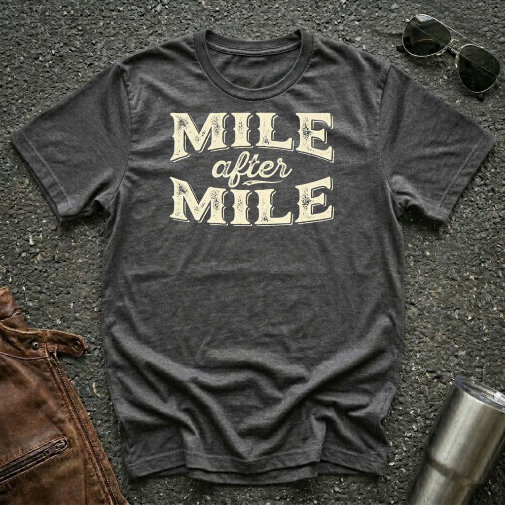 A dark heather graphic t-shirt laid flat on asphalt with sunglasses, a brown leather jacket, and a metal tumbler nearby. The shirt features the text 'MILE after MILE' in distressed cream lettering.