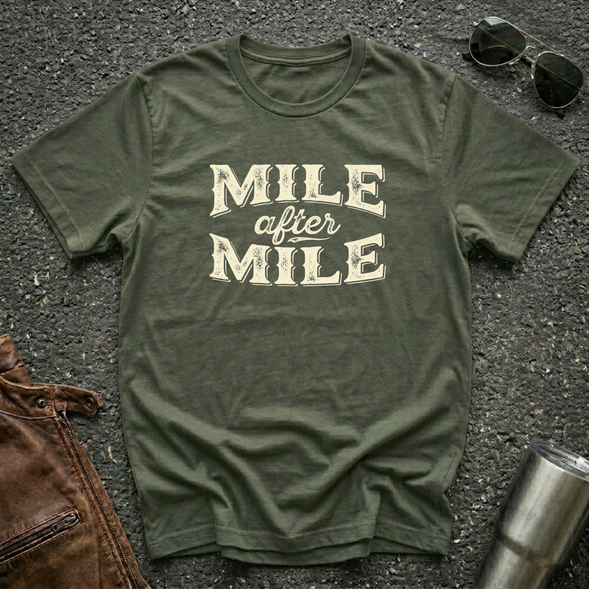 A military green graphic t-shirt laid flat on asphalt with sunglasses, a brown leather jacket, and a metal tumbler nearby. The shirt features the text 'MILE after MILE' in distressed cream lettering.