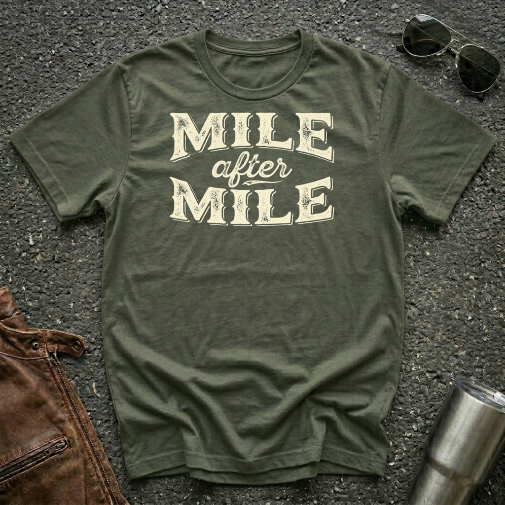 A military green graphic t-shirt laid flat on asphalt with sunglasses, a brown leather jacket, and a metal tumbler nearby. The shirt features the text 'MILE after MILE' in distressed cream lettering.